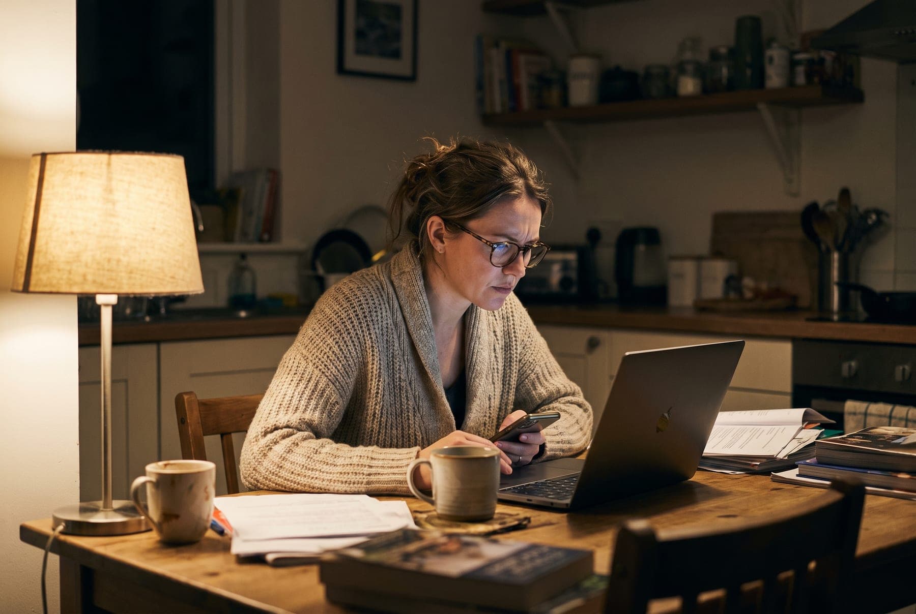 A woman at a kitchen table in the evening with a laptop and phone, organising